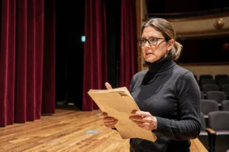 Metteure en scène de théâtre tenant un script face à une scène vide avec rideau rouge et sièges en velours en arrière-plan